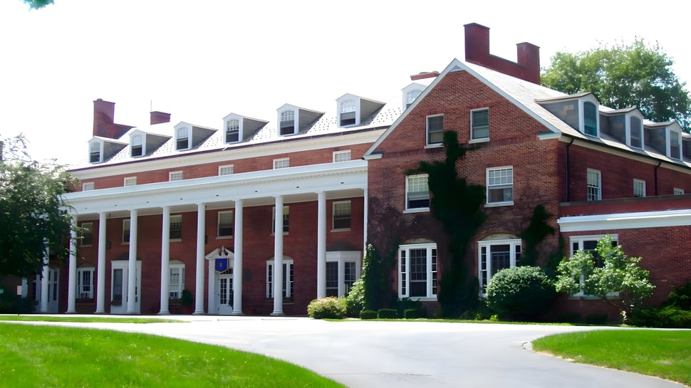 El gran edificio de ladrillo con pórtico y buhardillas se encuentra en el campus de la Miss Hall’s School en un prado con árboles.
