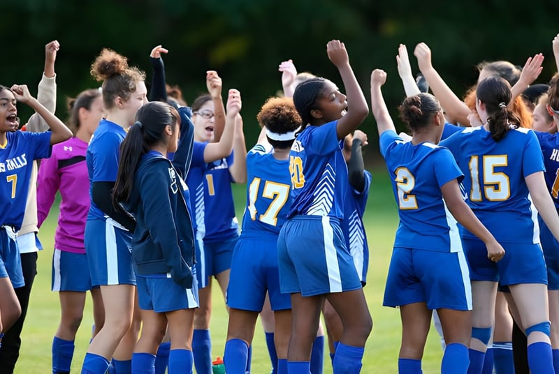 Un grupo de jugadoras de fútbol en camisetas azules en el campo de la Miss Hall’s School está con los brazos en alto.