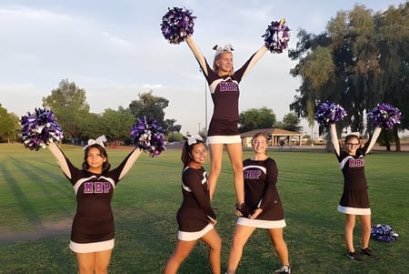 Estudiantes de la Mission Heights Preparatory High School realizan una actuación de cheerleading en un campo de césped con árboles de fondo.