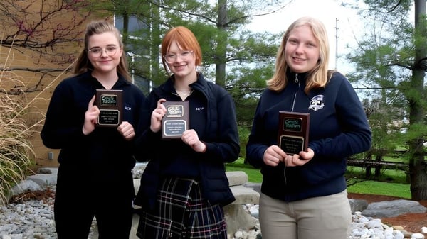 Tres estudiantes de la Mitchell District High School están con premios frente a una pared de piedra y árboles.