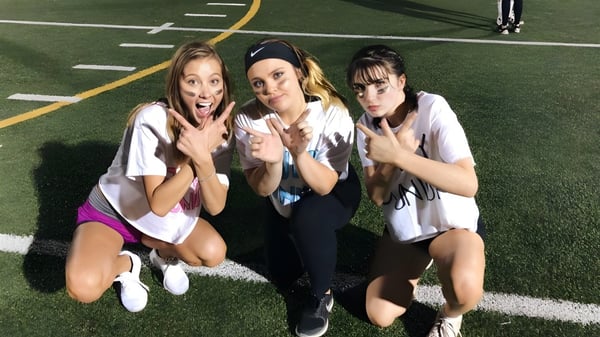 Tres estudiantes de la Monarch High School posan en el campo deportivo con marcas de campo y goles al fondo.