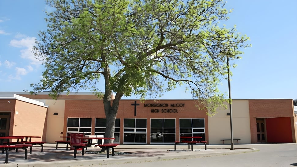 Un gran árbol con densa hoja verde está frente al edificio de ladrillo de la Monsignor McCoy High School.