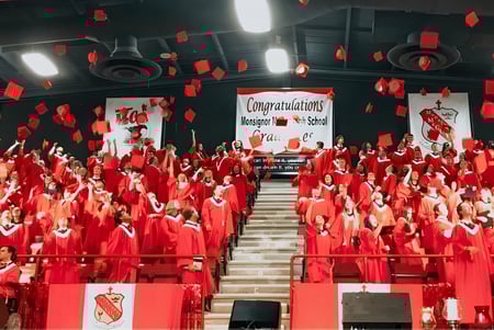 Estudiantes de la Monsignor McCoy High School llevan túnicas rojas en la ceremonia de graduación en un interior.