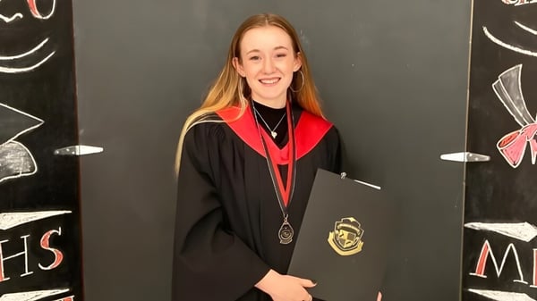 Una joven en toga de graduación se encuentra frente a una pizarra con dibujos en el campus de la Montague Regional High School.