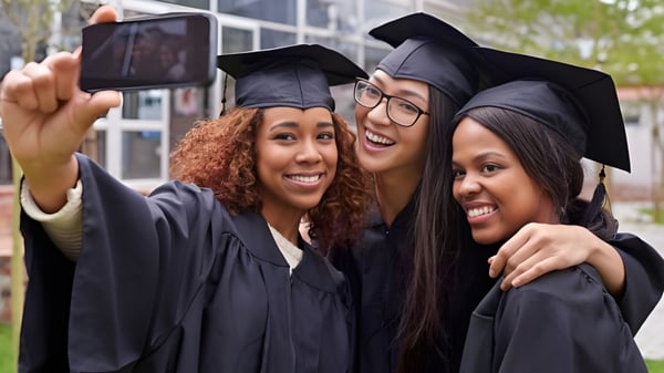 Tres graduadas de la Montcalm Secondary School posan en sus togas de graduación para un selfie frente a un edificio escolar con árboles.