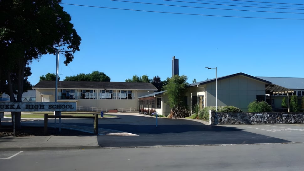 El edificio escolar con una alta torre de la Motueka High School se encuentra al fondo rodeado de árboles y un estacionamiento pavimentado.