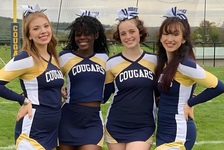 Cuatro estudiantes de la Mount Blue High School posan juntas en el campo deportivo con montañas de fondo.