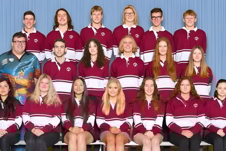 Un grupo de estudiantes de la Mount Compass Area School posan en uniformes escolares a rayas burdeos y blancos en el gimnasio.
