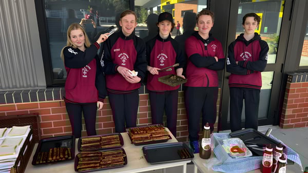 Cinco estudiantes de la Mount Compass Area School están de pie en uniformes burdeos uniformes frente a una tienda de comestibles y suministros.