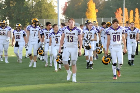 Un grupo de jugadores de fútbol en uniformes blancos de los Rams corre por el campo de la Mount Douglas Secondary School rodeado de árboles otoñales.