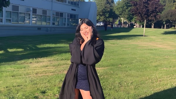 Una joven en un abrigo negro está de pie en una pradera frente al edificio de la Mount Douglas Secondary School.