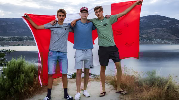 Tres estudiantes de la Mount Elizabeth Middle/Secondary School sostienen una gran bandera roja frente a un lago y un paisaje montañoso.