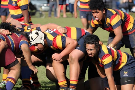 Un grupo de jugadores de rugby de la Mount Roskill Grammar está en un agrupamiento en un campo de césped con espectadores al fondo.