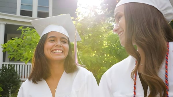 Dos estudiantes de la Mount Saint Michael están en sus togas de graduación frente a una casa con porche y plantas verdes.