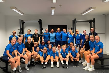 Un grupo de estudiantes de The Mount School York posan en uniformes azules en el gimnasio con equipos deportivos.