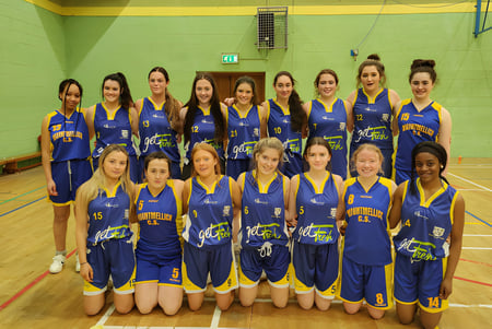 Un grupo de jóvenes jugadoras de baloncesto en camisetas azules y amarillas posan en el gimnasio de la Mountmellick Community School.