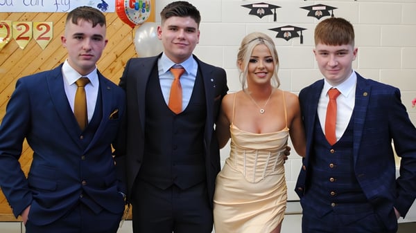 Cuatro jóvenes posan frente a una pared decorada con birretes de graduación en el campus del Moyne College.