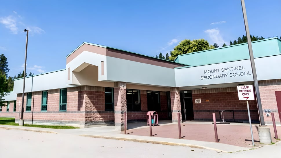 El edificio de ladrillo de varios pisos con techo verde de la Mt. Sentinel Secondary School está rodeado de árboles bajo un cielo despejado.
