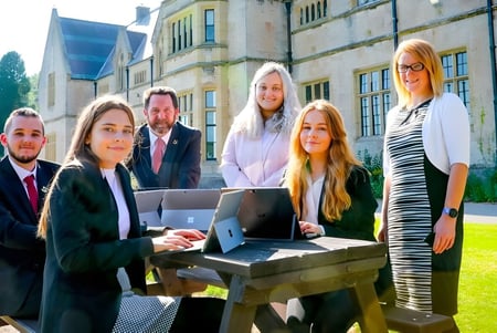 Un grupo de jóvenes en el Myddelton College está vestido de manera formal alrededor de un portátil sobre un pedestal de piedra frente a un edificio histórico con moderna cristalera.