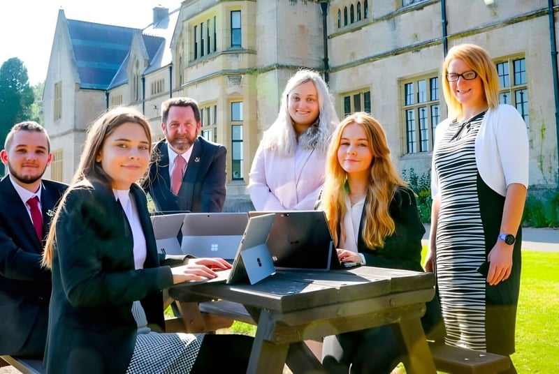 Un grupo de jóvenes en el Myddelton College está vestido de manera formal alrededor de un portátil sobre un pedestal de piedra frente a un edificio histórico con moderna cristalera.