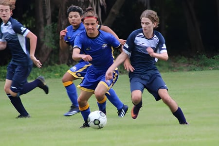 Estudiantes de Nambour State College juegan al fútbol en un campo de césped con árboles de fondo.