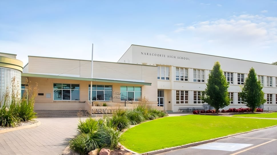 El moderno edificio escolar de varios pisos de la Naracoorte High School con césped bien cuidado y plantas en primer plano bajo un cielo azul.