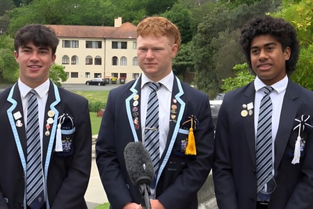 Tres alumnos del Nelson College for Boys están frente a un edificio con un fondo verde y boscoso.