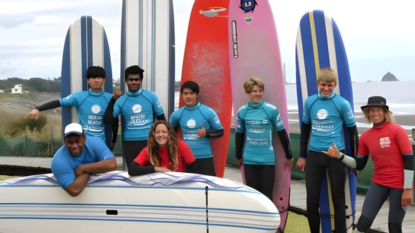 Estudiantes de la New Plymouth Boys' High School posan con tablas de surf frente a un paisaje costero con montañas al fondo.