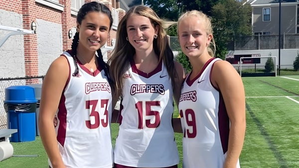 Tres estudiantes de la Newburyport High School están en la camiseta deportiva del equipo Clippers en un césped frente a un edificio de ladrillo.