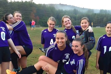 Un grupo de jugadoras de fútbol del Newcomb Central School District está en el césped de un campo con un bosque al fondo.