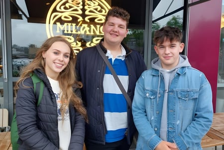Tres estudiantes de la Newtown School están juntos frente a un restaurante con un letrero decorativo.