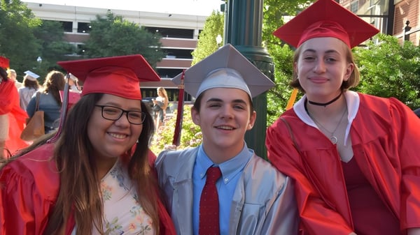Tres graduadas de la Niskayuna High School en togas rojas están frente a un edificio universitario.
