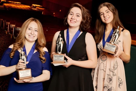 Tres estudiantes de la North Broward Preparatory School posan con premios frente a un telón de fondo rojo en el auditorio.