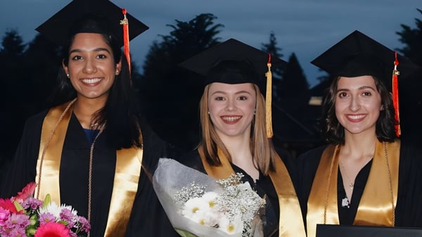 Tres graduados de la North Delta Secondary School en togas de graduación están juntos frente a árboles y un cielo oscuro.