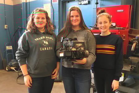 Tres estudiantes están juntas en un taller de la North Lambton Secondary School con herramientas de fondo.