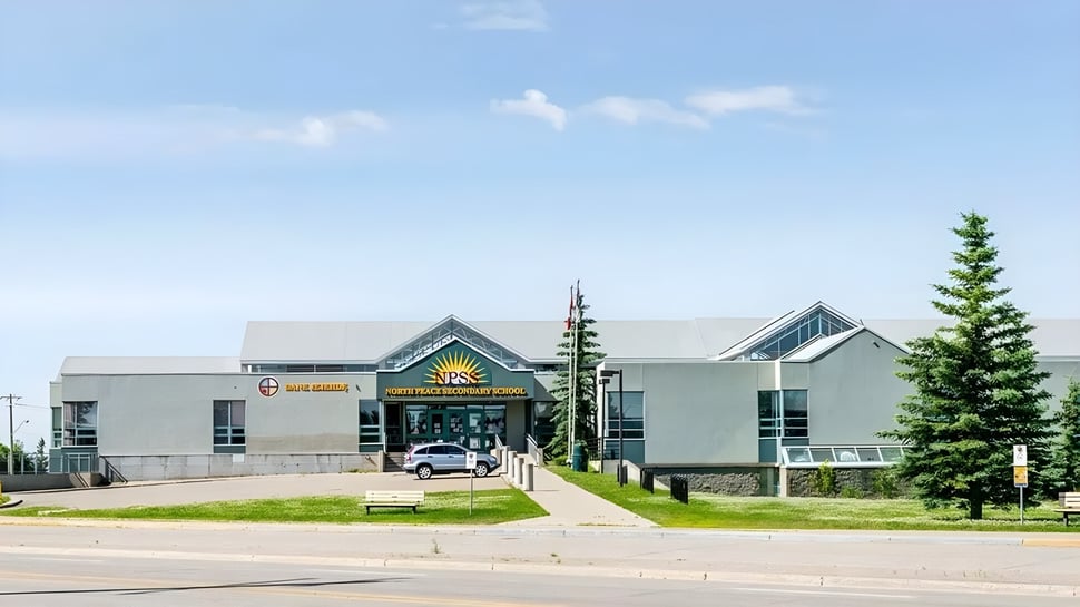 El moderno edificio principal de la North Peace Secondary School se encuentra frente a un prado verde con árboles bajo un cielo azul.