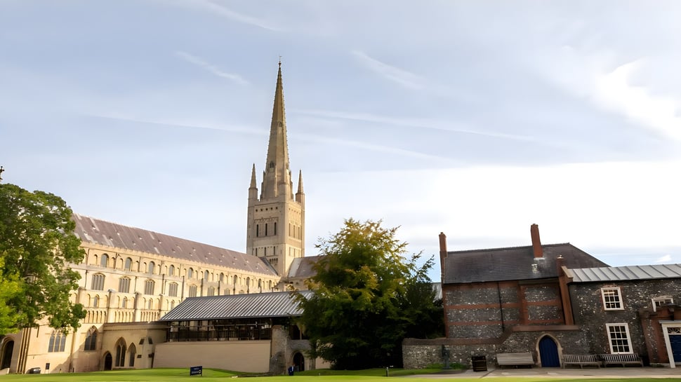 La alta torre de una catedral sobresale de los edificios en el terreno de la Norwich School bajo un cielo nublado.