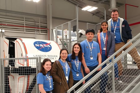 Un grupo de estudiantes de la Norwood International High School está en uniformes de la NASA frente a un gran logo de la NASA y un modelo de Lander Rover.