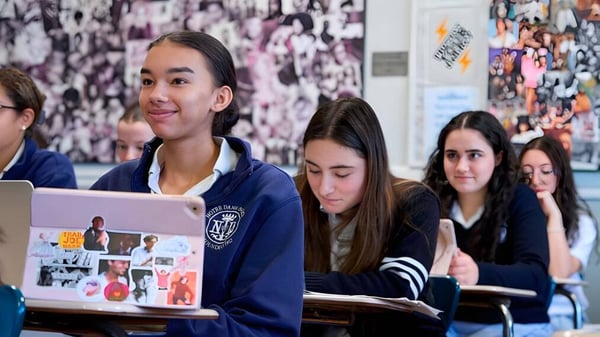 Un grupo de estudiantes está sentado en el aula de Notre Dame High School con un collage de imágenes y carteles en el fondo.