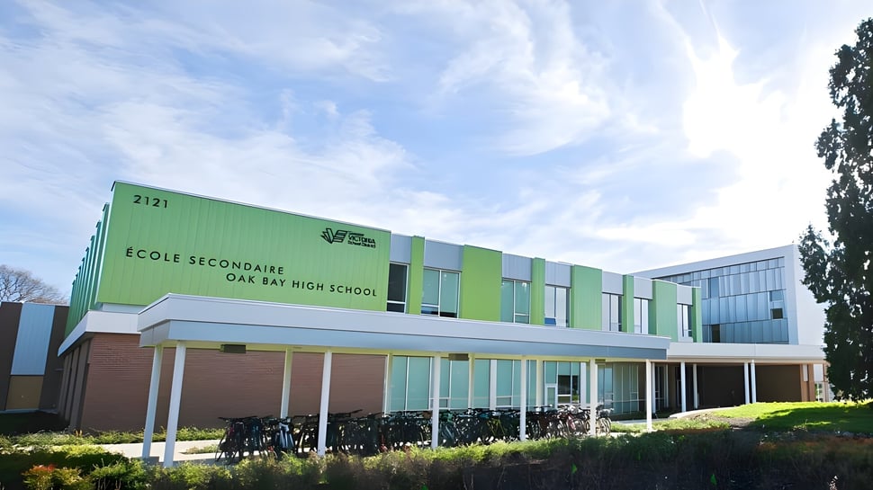 El moderno edificio escolar de varios pisos de la Oak Bay Secondary High School con fachada verde y grandes ventanas está rodeado de árboles.