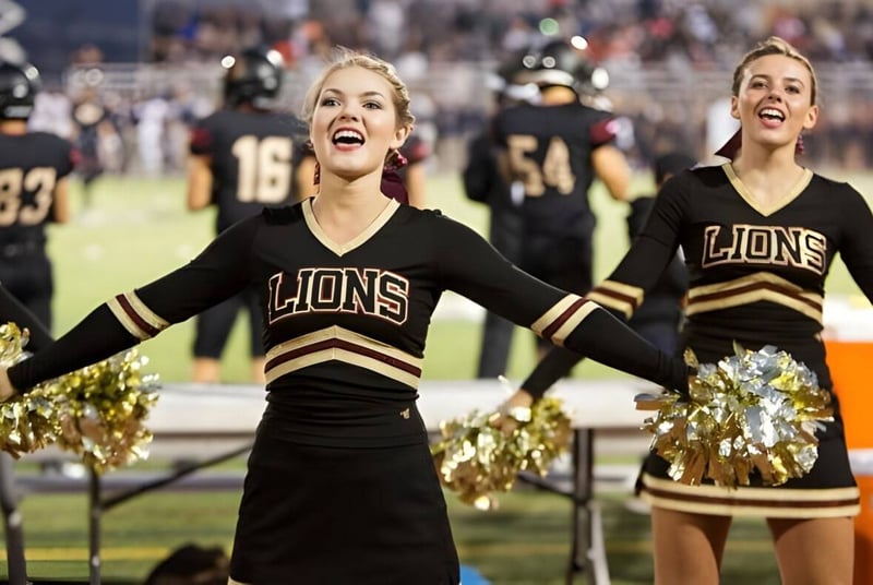 Dos animadoras de la Oaks Christian School en uniformes negro-dorados animan al borde del campo de un partido de fútbol americano.