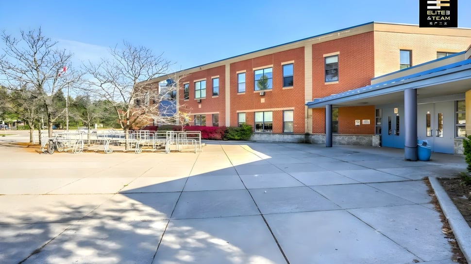 El edificio de ladrillo de la Oakville Trafalgar High School con acentos azules y rojos se encuentra detrás de un patio pavimentado y árboles.