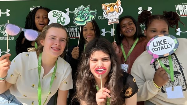 Un grupo de estudiantes sonrientes posa frente a una pared de fotos con gráficos en el campus de la Oldfields School.