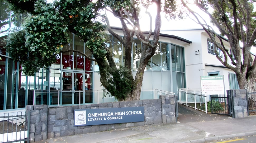 El cartel de piedra de la Onehunga High School está frente a un edificio moderno y rodeado de vegetación verde.