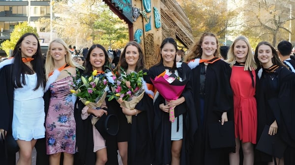 Un grupo de egresadas de la Onehunga High School está frente a un mural colorido sosteniendo ramos de flores.