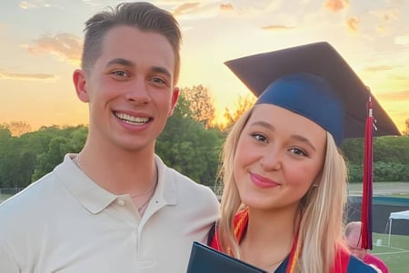Dos graduados de la Orono High School en ropa académica frente a un paisaje pintoresco con árboles y un atardecer.