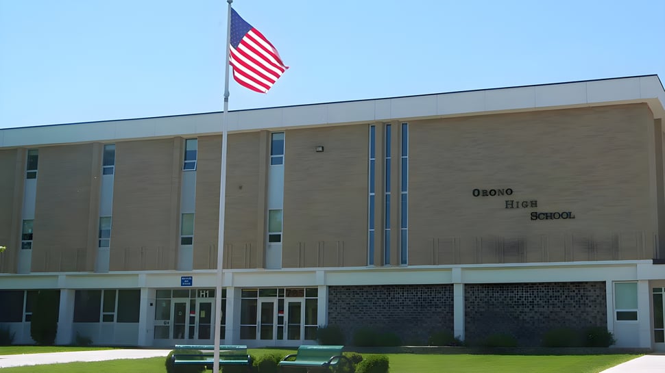 El gran edificio de ladrillo de la Orono High School con una bandera americana en el techo y un césped con bancos delante.