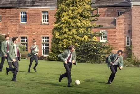 Un grupo de personas vestidas de manera informal camina por un campo de hierba frente a un edificio de ladrillo en el terreno de la Oswestry School.