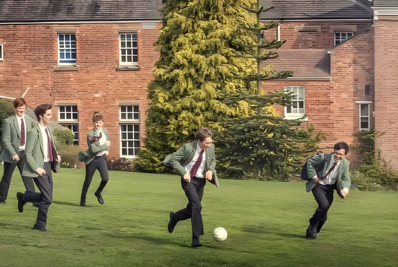 Un grupo de personas vestidas de manera informal camina por un campo de hierba frente a un edificio de ladrillo en el terreno de la Oswestry School.