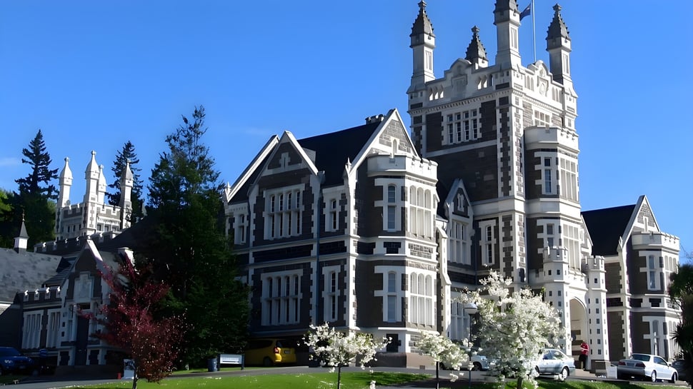 El edificio principal gótico de la Otago Boys' High School está rodeado de árboles en flor bajo un cielo despejado.